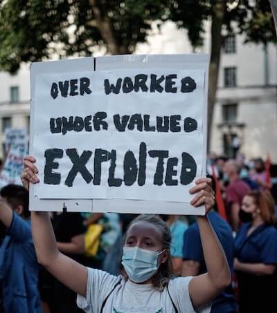 A person at a rally with a mask on holding a sign that says "over worked under valued exploited."