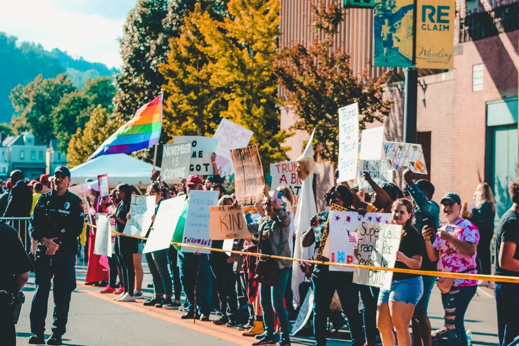 People lined up on a sidewalk behind a tape holding various signs and one rainbow flag.