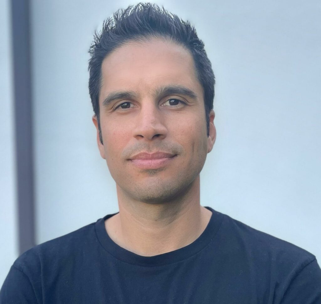 Zoltan Csaplar in a dark blue t-shirt standing in front of a white wall.